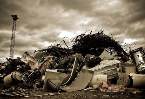 A large open lot in Mottingham features an extensive pile of mixed construction and industrial waste, including metal sheets, plastic piping, and broken appliance parts, all arranged in a chaotic heap. The materials vary in colour, with white, grey, and metallic tones dominating, and textures ranging from smooth and reflective metal surfaces to rough, corroded, and weathered elements. In the background, a tall, slender telecommunications tower and a cloudy, overcast sky create a stark contrast against the cluttered foreground. The waste appears to have been recently collected, with some items leaning on others or partially buried beneath debris, indicative of a removal operation. Surrounding the scrap heap is a paved surface typical of a commercial or industrial yard in the vicinity of London suburbs, possibly Nottingham. The scene is captured in natural light, with diffused sunlight filtering through the clouds, evoking a neutral, observational tone that underscores the need for professional rubbish removal services such as those provided by Commercial Waste Removal Mottingham to manage and clear such waste efficiently and safely.