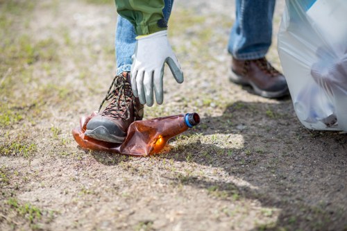 Close-up of a person wearing a white work glove, placing a brown glass bottle on the ground covered with dirt and small patches of grass. The individual is dressed in blue jeans and dark work boots, with part of a white plastic bag visible nearby. The setting appears to be an outdoor area, possibly a parking space or driveway with natural light illuminating the scene, indicating a cleanup or rubbish disposal activity. The focus is on the bottle and hand, highlighting waste collection as part of rubbish removal services offered by Commercial Waste Removal Mottingham, serving local areas near the postcode in Mottingham, London. The environment looks clean but shows typical outdoor ground conditions associated with waste management tasks, emphasizing the importance of responsible rubbish disposal and recycling practices involving glass bottles.