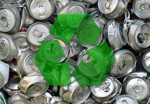 A close-up view of a large pile of empty, silver aluminium beverage cans stacked haphazardly outdoors, with some cans showing visible pull-tab openings and embossed markings on the tops. The cans have smooth, shiny surfaces that reflect light, with a mix of slightly crumpled and intact cans. Positioned centrally over the cans is a large, green recycling symbol composed of three arrows forming a triangle, indicating environmentally conscious disposal or recycling efforts. The background consists of more cans filling the frame, suggesting a collection point or waste material ready for removal. This scene is representative of waste management services offered by Commercial Waste Removal Mottingham, focusing on rubbish collection and recycling in the local area, possibly near residential or commercial premises in the vicinity of Nottingham or the postcode region associated with Mottingham.