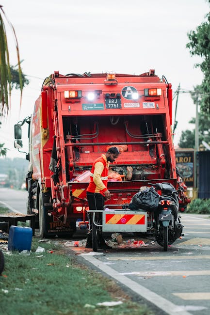 A modern white waste collection vehicle with a large, partially open rear compactor section, positioned on a narrow urban street with cobblestone paving. The vehicle's rear is designed for garbage com
