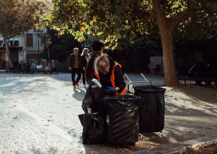 A woman wearing a high-visibility orange vest and blue gloves is actively collecting waste and emptying a black rubbish bag into a large black trash bin on a paved outdoor area. The bin is positioned 