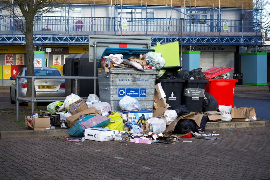 A woman wearing a high-visibility orange vest and blue gloves is actively collecting waste and emptying a black rubbish bag into a large black trash bin on a paved outdoor area. The bin is positioned 