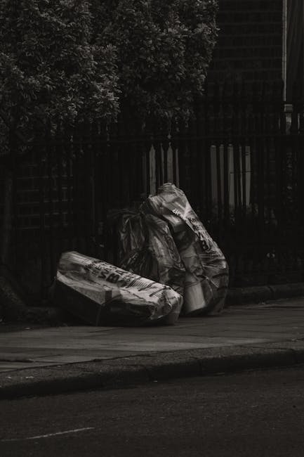 Two large black rubbish bags, made of durable plastic with a slightly crinkled and shiny surface, are positioned on a sidewalk near the curb. The bags appear to be filled with waste materials, causing