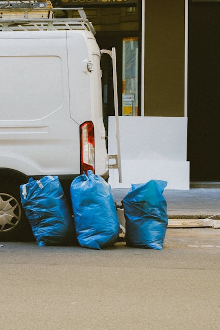 A sanitation worker in a yellow and red uniform is using a tool at the back of a large red rubbish collection truck, which is stationed on the side of a roadway. The truck's rear panel is open, reveal